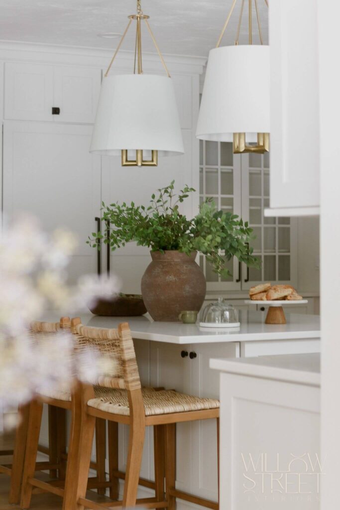 Kitchens tend to carry a lot of emotional weight — they’re gathering spots, coffee stops, morning chaos stations, and the place you stand when you can’t decide what you want but open the fridge anyway. So when we were designing this space, we knew it had to feel easy, but also grounded. Comfortable, but quietly elevated. And yes, beautiful from every angle.
This kitchen is part of a new construction home, which gave us the freedom (and the blank canvas) to play with contrast and softness in equal measure. The palette leans into warm whites and layered neutrals — think sun-washed walls and white oak floors underfoot — but it’s not shy. Deep black accents and bold brash details cut through the calm, anchoring the space with just the right amount of edge.